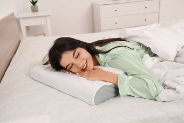 Pijamalı genç bir kadın evdeki rahat yatakta yumuşak ortopedik yastığa uzanıyor.