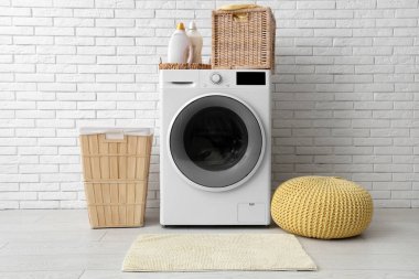 Beyaz tuğla duvarın yanında sepetleri, puf ve deterjanı olan modern çamaşır makinesi.
