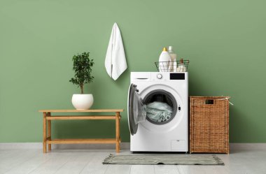 Yeşil duvarın yanında yedek kulübesi, deterjanı ve sepeti olan modern çamaşır makinesi.