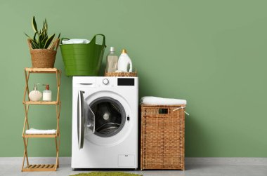 Yeşil duvarın yanında rafları, sepetleri ve deterjanları olan modern çamaşır makinesi.