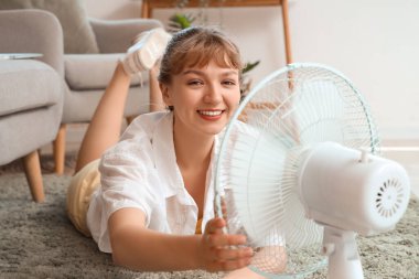 Evde yerde yatan, elinde vantilatör olan genç bir kadın.