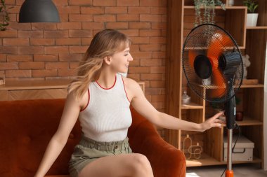 Evdeki koltukta elektrikli yelpaze çalan genç bir kadın oturuyor.