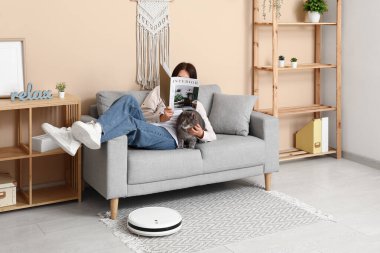 Kedi ve robot elektrikli süpürgeyle kanepede dinlenen genç bir kadın.