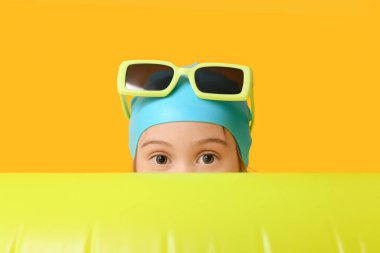 Şişme yüzüklü, yüzme şapkalı ve sarı arka planda güneş gözlüklü şirin bir kız.