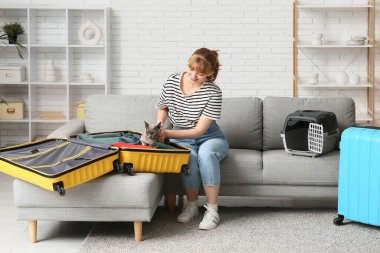 Evdeki kanepede oturan sevimli kedisi, hayvan taşıyıcısı ve bavulu olan genç bir kadın.