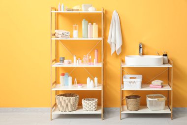 Banyoda sarı duvarın yanında lavabo ve banyo malzemeleri olan raflar.