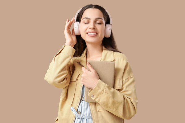 Красивая молодая женщина в наушниках слушает аудиокнигу и держит книгу на коричневом фоне