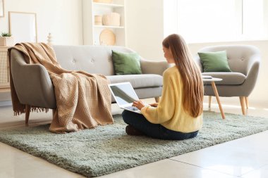 Genç bir kadın dizüstü bilgisayar kullanıyor ve oturma odasında yerde oturuyor.