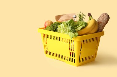 Farklı taze ürünlerle dolu sarı alışveriş sepeti ve bej arka planda elmalar