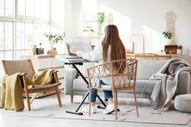 Elinde not kağıtları olan genç bir kadın oturma odasında, arka planda sentezleyici oynuyor.