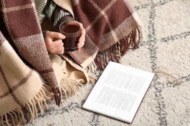 Ekose kumaş ve bir fincan sıcak çikolatayla evde yerde kitap okuyan güzel genç bir kadın.