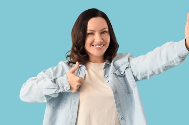 Güzel, olgun, mutlu bir kadın mavi arka planda selfie çekerken başparmak hareketi yapıyor.