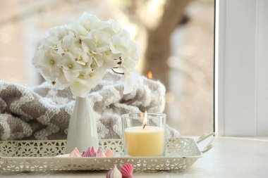 Çiçekli vazo, yanan mum ve pencerenin yanındaki tepside kremalar, yakın plan.