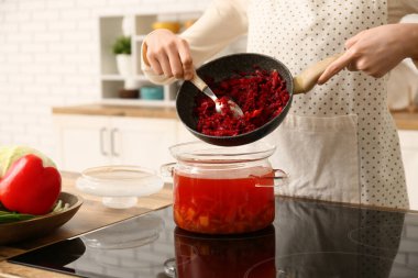 Genç bir kadın mutfakta lezzetli borş ile tencereye sebze kızartma ekliyor.