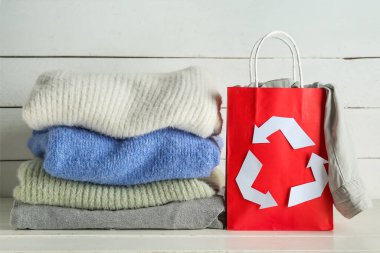 Geri dönüşüm amblemi ve beyaz ahşap arka planda giysileri olan alışveriş torbası.