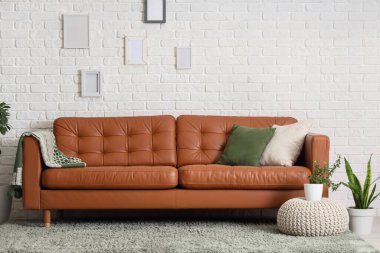 Beyaz tuğla duvarın yanında şık deri kanepe, kanepe ve ev bitkileriyle oturma odasının içinde.