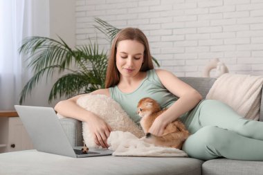Evdeki kanepede dizüstü bilgisayarı ve sevimli kedisi olan genç bir kadın yatıyor.