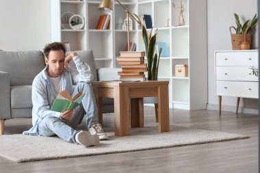 Genç adam evde yerde kitap okuyor.