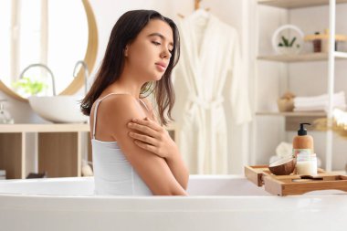 Güzel genç bir kadın banyoda vücuduna hindistan cevizi yağı sürüyor.