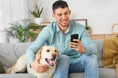 Cep telefonu ve evdeki koltukta oturan Labrador köpeği olan genç adam.