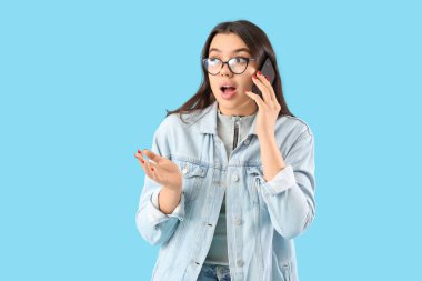 Gözlüklü genç kadın mavi arka planda cep telefonuyla konuşuyor.