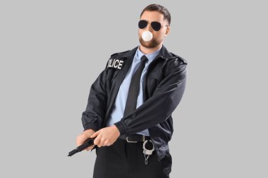 Gri arka planda sakız sallayan silahlı bir erkek polis memuru.