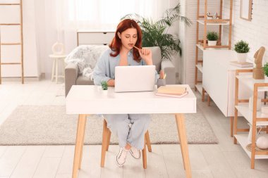 Evdeki masada dizüstü bilgisayar kullanan düşünceli genç bir kadın.