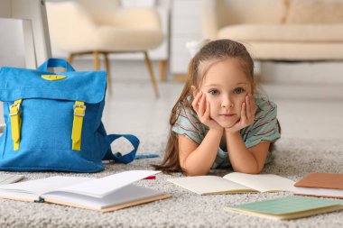 Not defterleri ve sırt çantasıyla evde yerde yatan sevimli küçük kız.