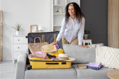 Genç Afro-Amerikalı kadın otel odasındaki kanepede seyahat aksesuarlarıyla bavulunu boşaltıyor.