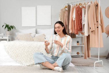 Yatak odasında giysileri olan cep telefonu kullanan genç bir kadın.