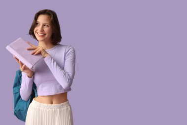 Sırt çantası ve eflatun arkaplan üzerine kitabı olan güzel bir kız öğrenci.