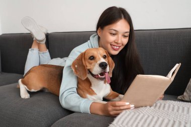 Genç bir kadın kitap okuyor ve oturma odasındaki kanepede sevimli bir Beagle köpeğiyle yatıyor.