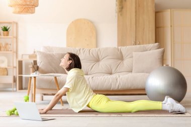 Dizüstü bilgisayarlı güzel bir kadın oturma odasında yoga yapıyor.