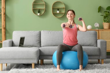 Oturma odasında halterli güzel bir genç bayan antrenman yapıyor.