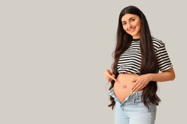 Güzel genç hamile bir kadın, gri arka planda karnı üzerine çizilmiş kalbi işaret ediyor.