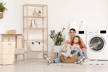 Genç baba ve kızı çamaşır sepetiyle banyoda çamaşır makinelerinin yanında oturuyorlar.