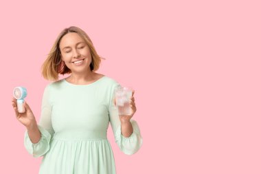 Mini elektrikli vantilatörü ve pembe arka planda bir bardak soğuk suyu olan olgun, mutlu bir kadın.