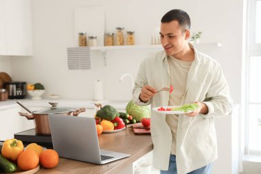Bilgisayarlı genç adam mutfakta pişmiş yemek yiyor.
