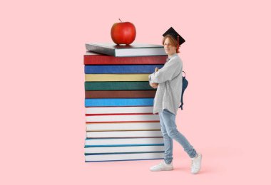 Mezuniyet şapkalı erkek öğrenci, bir sürü büyük kitap ve pembe arka planda elma.