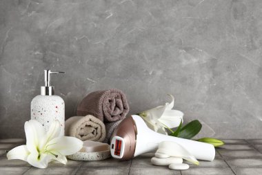 Gri duvarın yanındaki fayans masasında banyo malzemeleri ve nilüfer çiçekleri olan modern fotoepilatör.