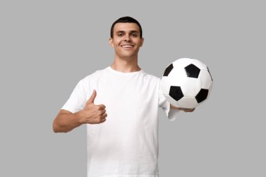 Futbol topuyla yakışıklı genç adam gri arka planda baş parmak hareketi gösteriyor.