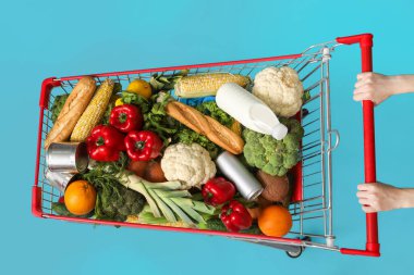 Mavi arka planda yiyecek dolu alışveriş arabasını iten kadın.