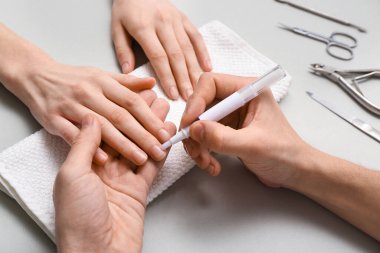 Erkek manikür ustası gri arka plandaki kadın tırnaklarına tırnak altı yağı sürüyor.