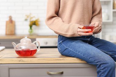 Çaydanlığın yanındaki masada oturan ve elinde bir fincan çay tutan genç kadın.