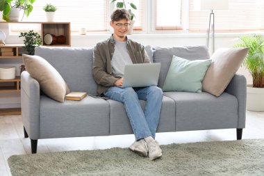 Bilgisayarlı erkek öğrenci evdeki kanepede ders çalışıyor.