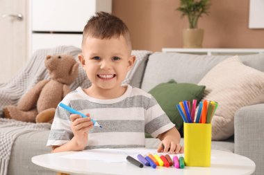 Evdeki masada keçeli kalemlerle resim çizen sevimli küçük çocuk.
