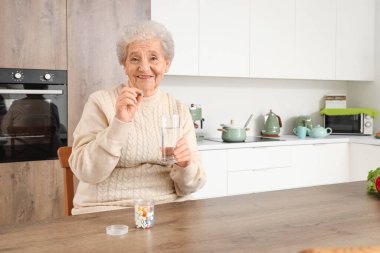 Bir bardak suyla mutfakta hap alan yaşlı bir kadın.