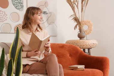 Genç, güzel bir kadın kitap okuyor ve beyaz oturma odasındaki kahverengi koltukta oturuyor.