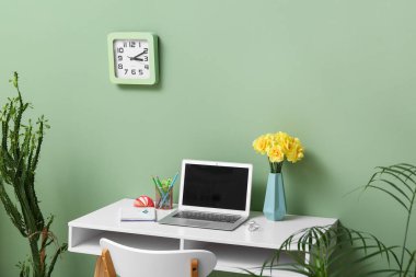 Bir buket narsisli çiçek, ev bitkileri ve dizüstü bilgisayarlı iş yeri şık bir ofiste yeşil duvarın yanında.