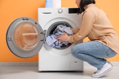 Young woman putting dirty clothes into washing machine on orange background